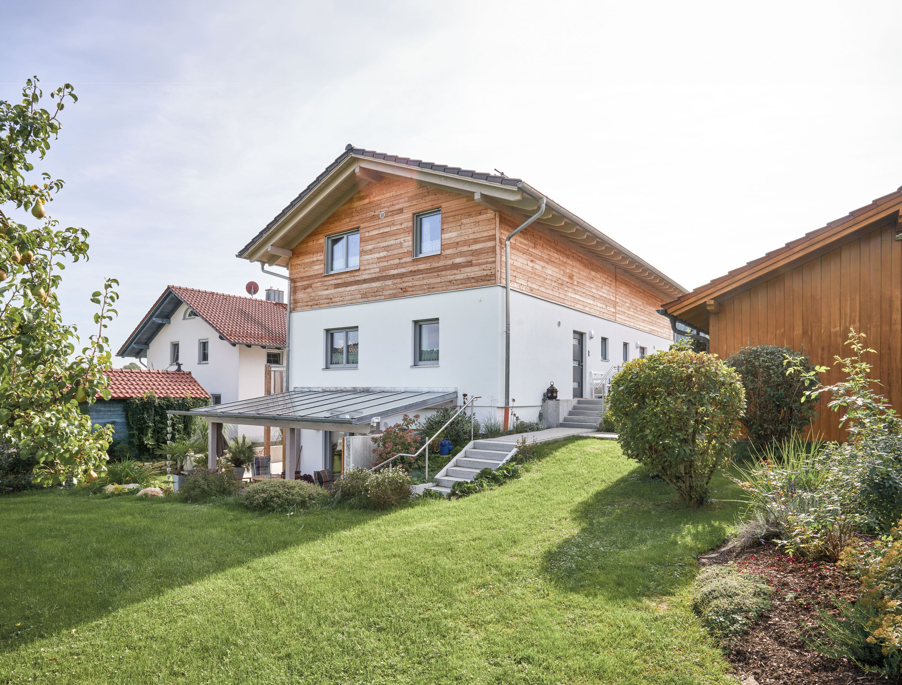 Zu sehen ist ein Haus mit einer zusätzlichen, barrierefreien Wohnung. Die Hausfassade ist weiß, im Dachgeschoss sind die Wände mit Lärchenbohlen zu sehen.