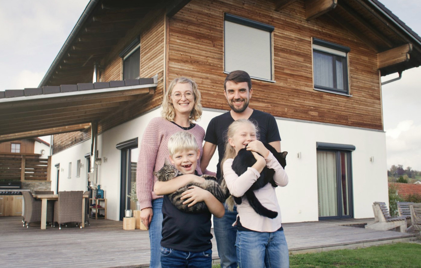 Zu sehen ist eine junge Familie vor ihrem Massivholzhaus, welches von der Firma Sonnleitner gebaut wurde.